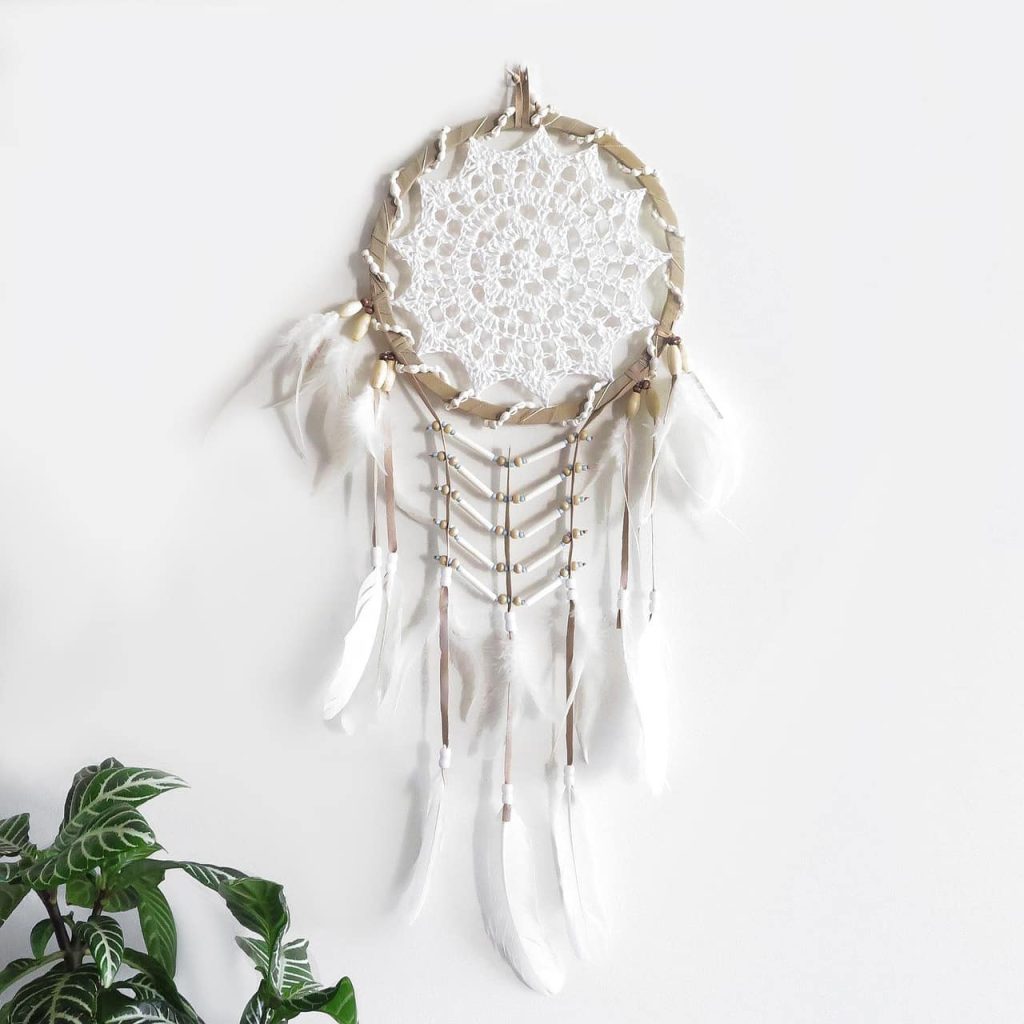 Attrape-rêves amérindien blanc avec napperon en dentelle, plumes légères et perles sur fond mural épuré avec plante verte