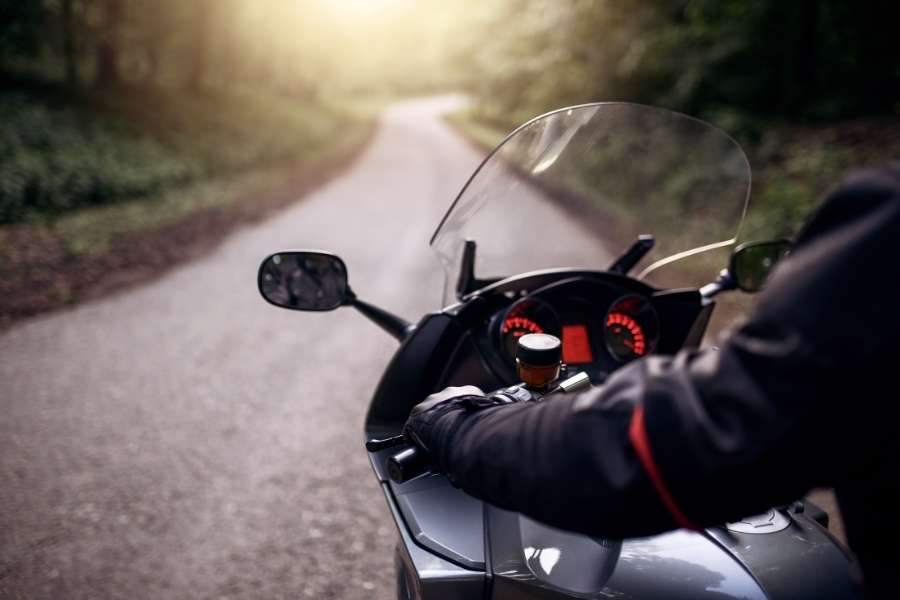 Vue subjective d'un motard gantés aux commandes d'une moto GT sur une route forestière sinueuse baignée de lumière, parfaite pour de longues sorties moto.