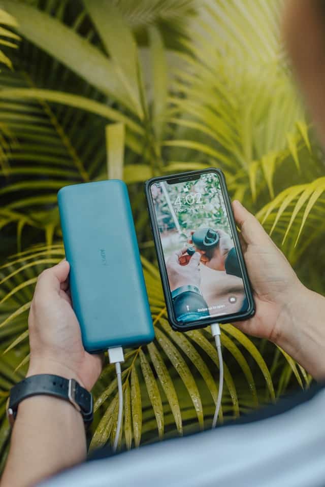 Une personne tenant un smartphone iPhone en charge relié à une powerbank bleue de marque Aukey sur un fond de feuillage tropical.