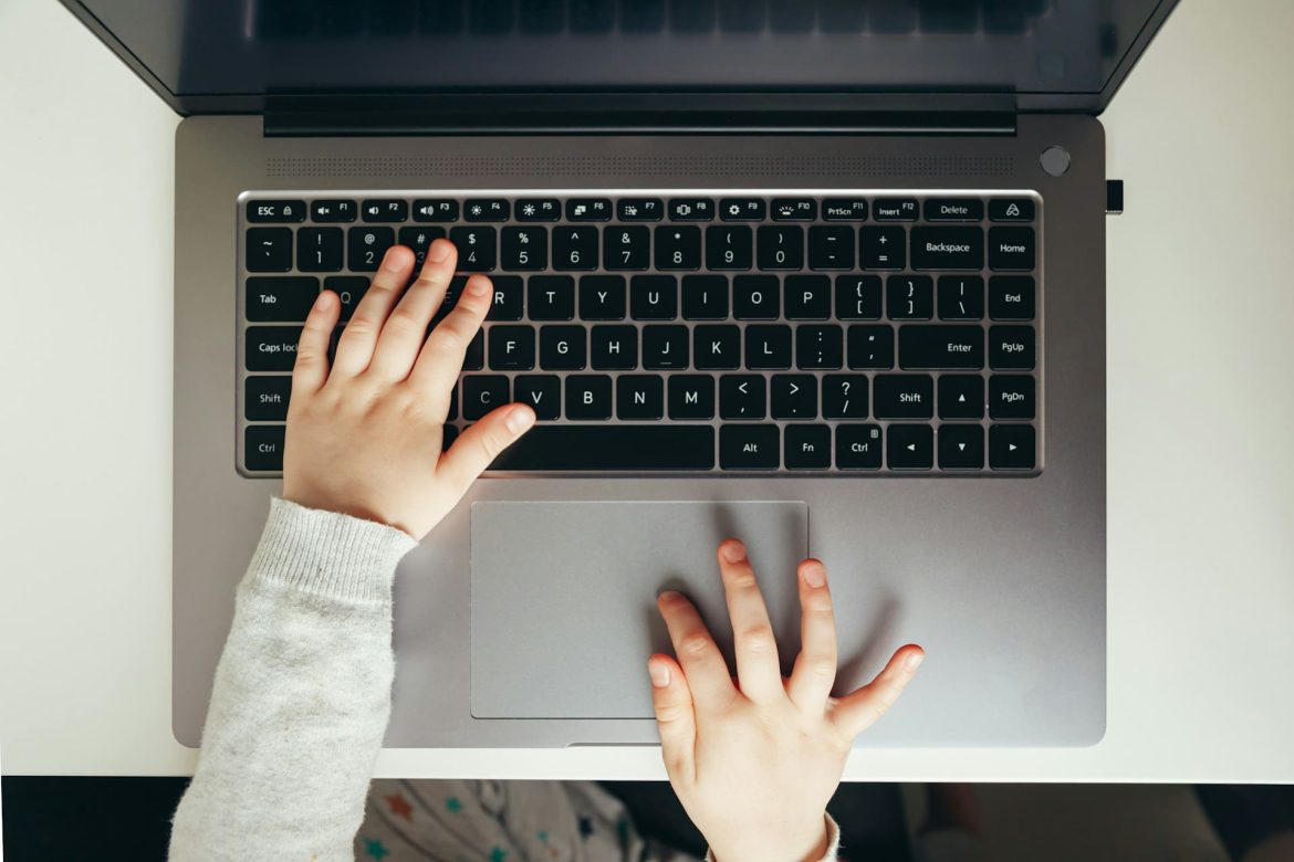 Vue de dessus des mains d'un jeune enfant posées sur le clavier et le pavé tactile d'un ordinateur portable, illustrant comment l'intelligence artificielle révolutionne l'apprentissage dès le plus jeune âge.