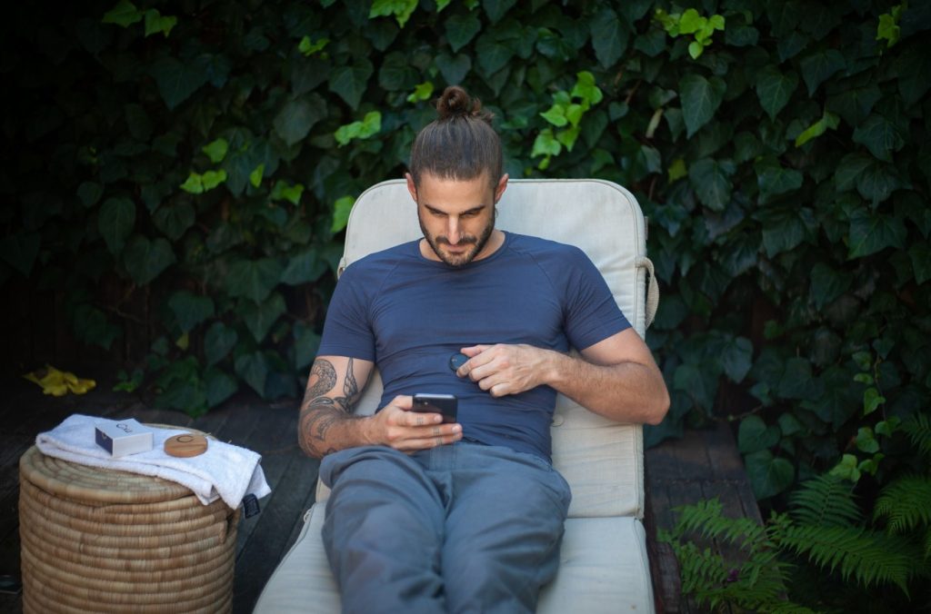 Un homme avec un chignon et des tatouages utilise un capteur de santé connecté sur sa poitrine tout en consultant son smartphone, illustrant les innovations IA 2026 en matière de bien-être.