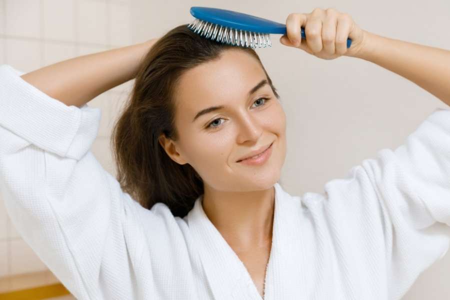 Une jeune femme souriante en peignoir blanc se brosse les cheveux avec une brosse à cheveux bleue.