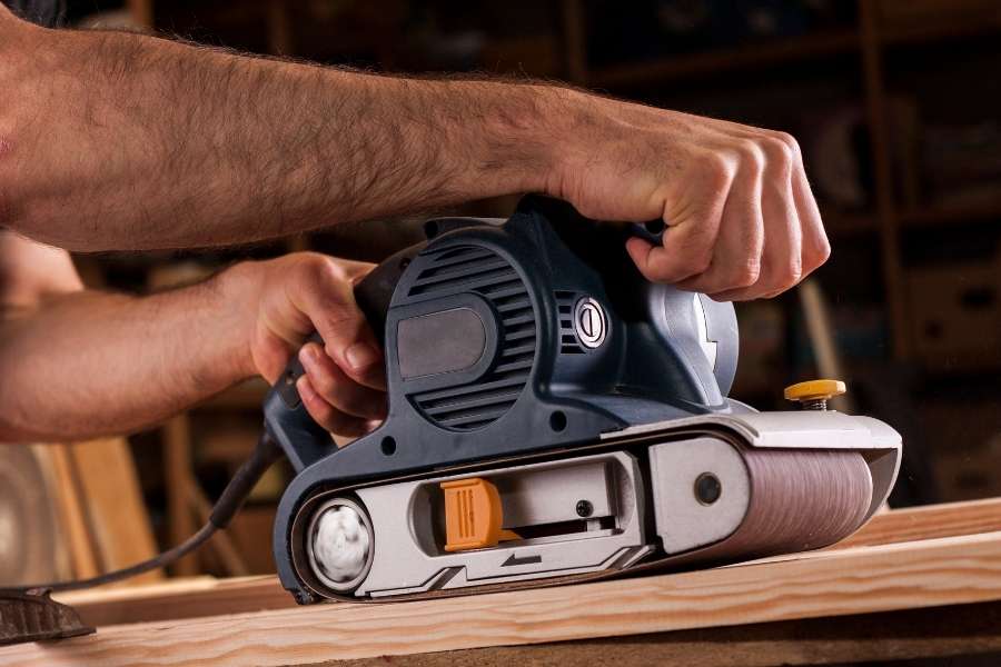 Gros plan sur les mains d'un artisan utilisant une ponceuse à bande électrique pour travailler une planche de bois.